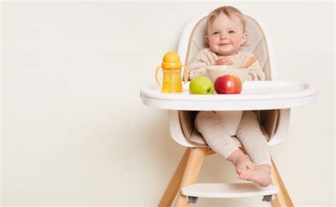 Kūdikis sėdi maitinimo kėdutėje ir bando valgyti iš šaukštelio